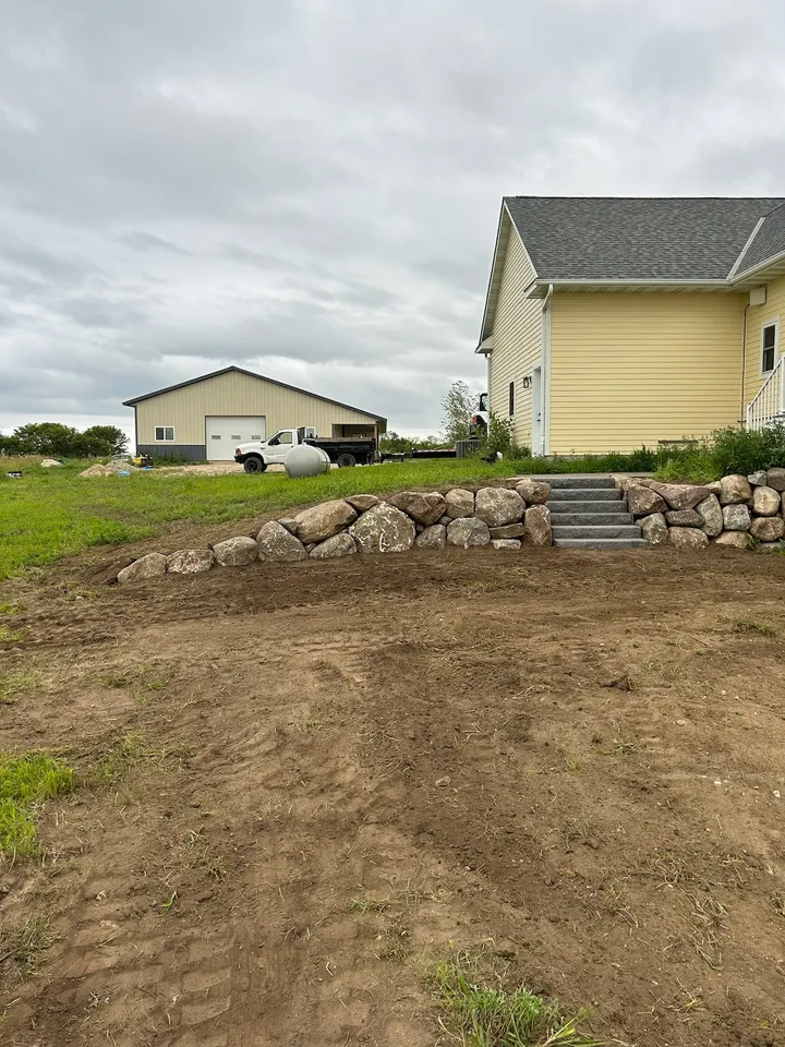 Low boulder retaining wall with stone steps in a front yard, house and detached garage behind, lawn not yet seeded