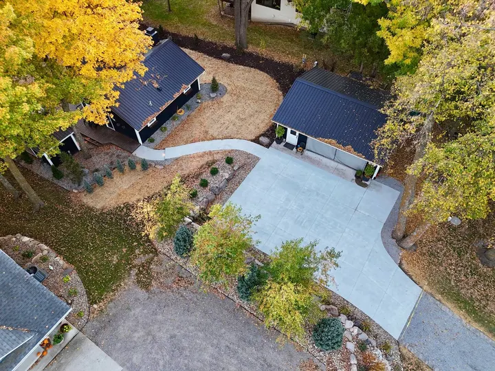 Drone view of a dark cabin and matching garage with a new concrete parking pad, a curved concrete walkway to the deck, boulder-edged planting beds, and straw-covered seeded lawn under fall color
