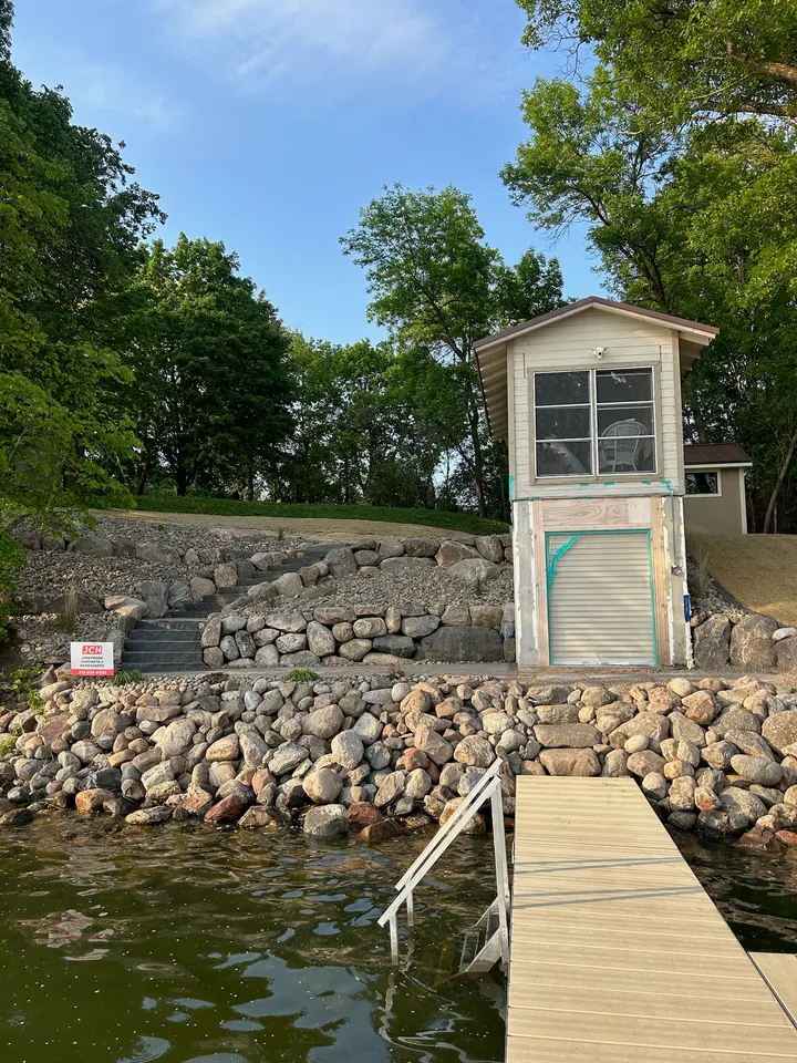 Fieldstone riprap bank below a tall narrow boathouse, with boulder steps climbing the slope on the left and a dock in the foreground.