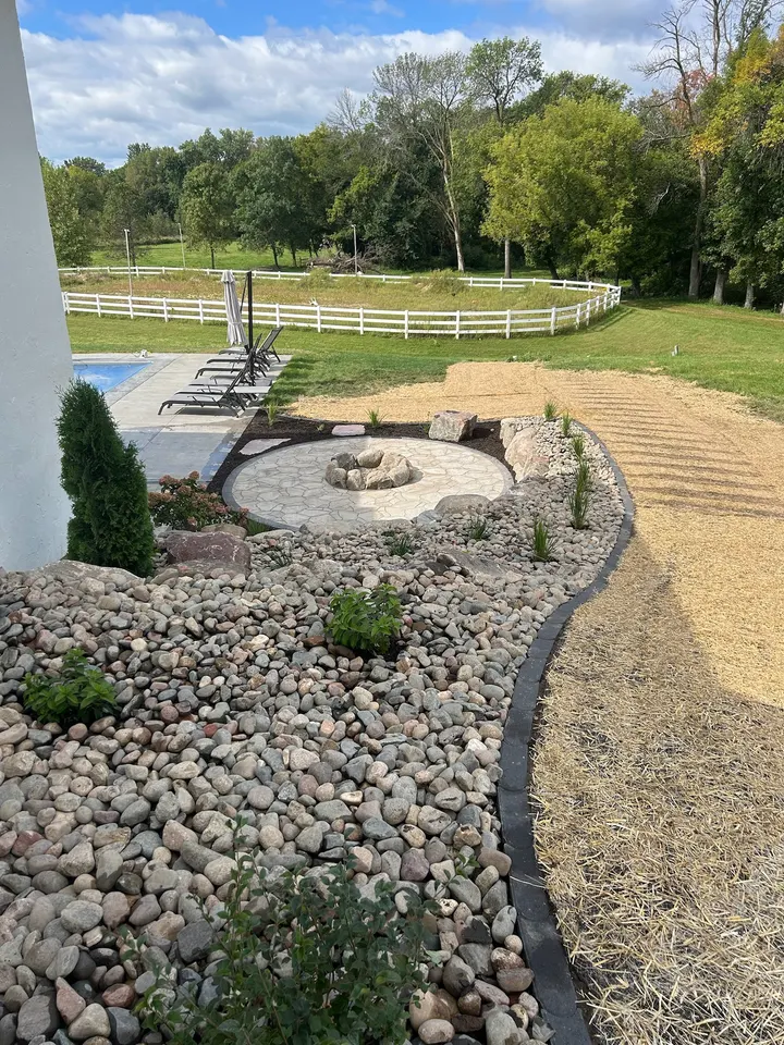 Round paver fire pit pad with a boulder ring, river rock beds with young plantings, and a pool with lounge chairs beyond, before any lighting was added