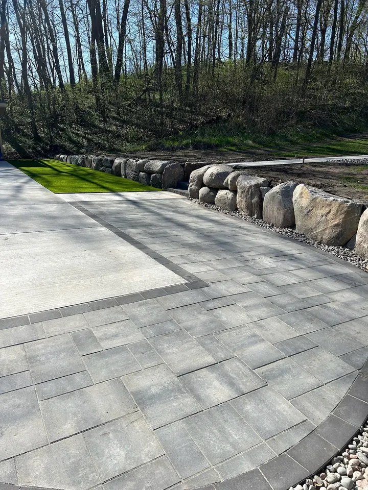 Dark gray paver field with a contrasting soldier-course border meeting a broom-finish concrete slab, with a boulder retaining wall holding the bank behind
