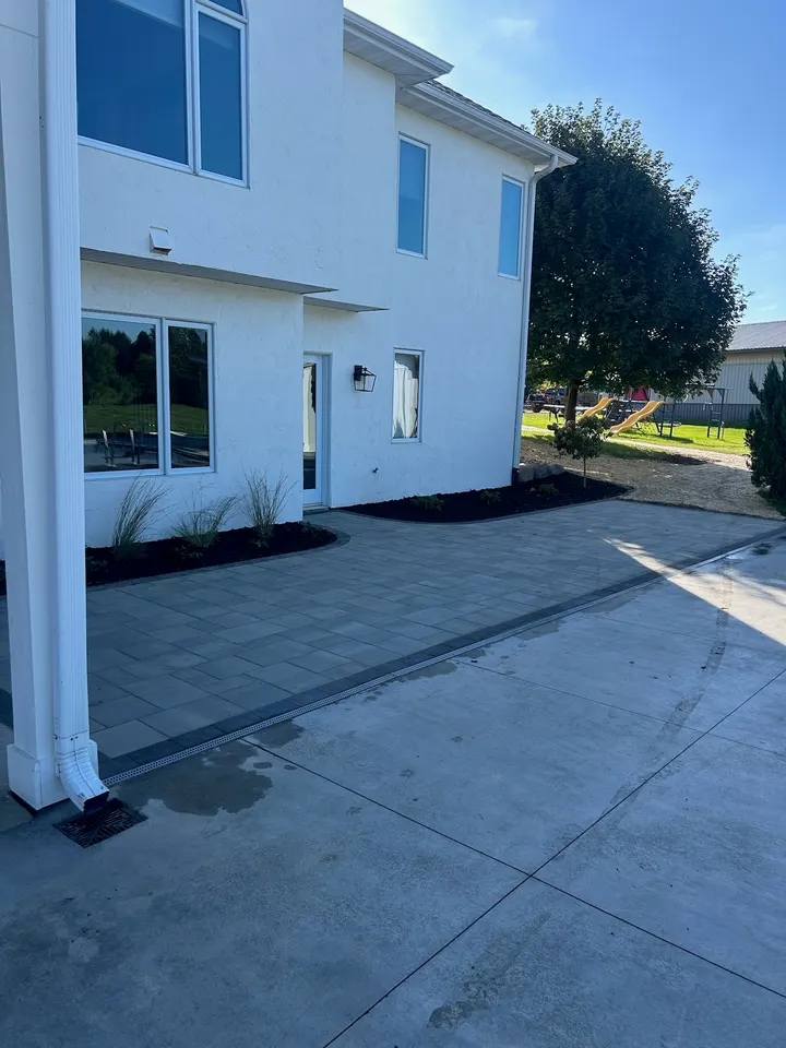 Paver patio laid tight to a white two-story home, with a mulched planting bed along the foundation and a downspout run to the edge