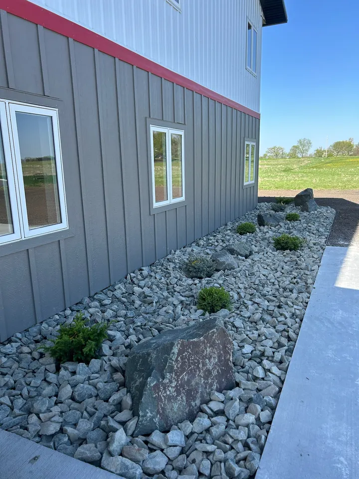 Gray river rock bed with small shrubs and a few dark boulders along the foundation of a gray steel-sided building, beside a concrete walk.