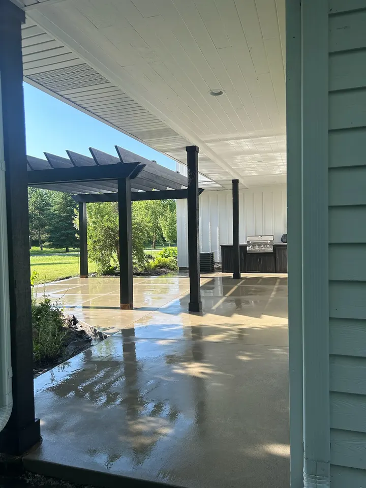Covered patio with a sealed concrete floor, dark posts, a pergola beyond, and a built-in grill station against the back wall